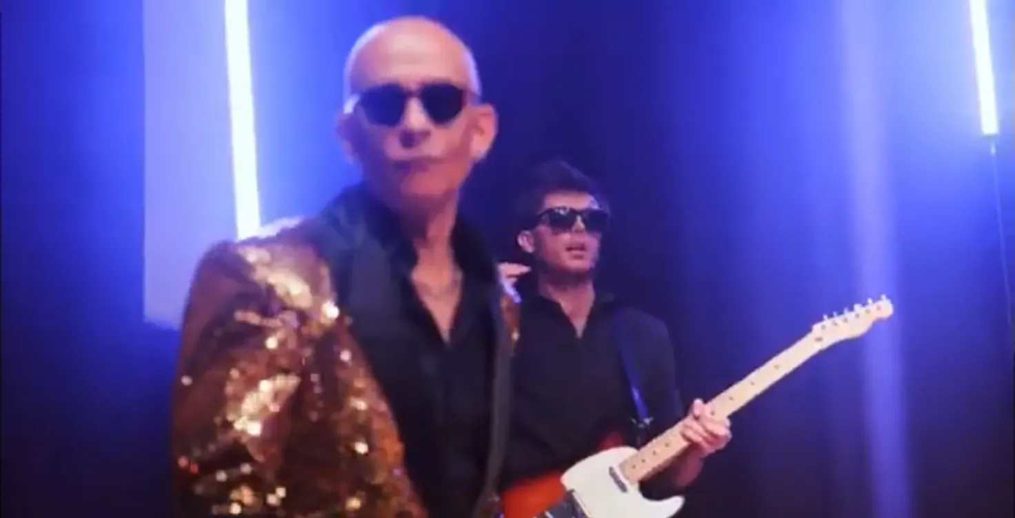 A guitarist in a gold sequin jacket and dark glasses under blue light
